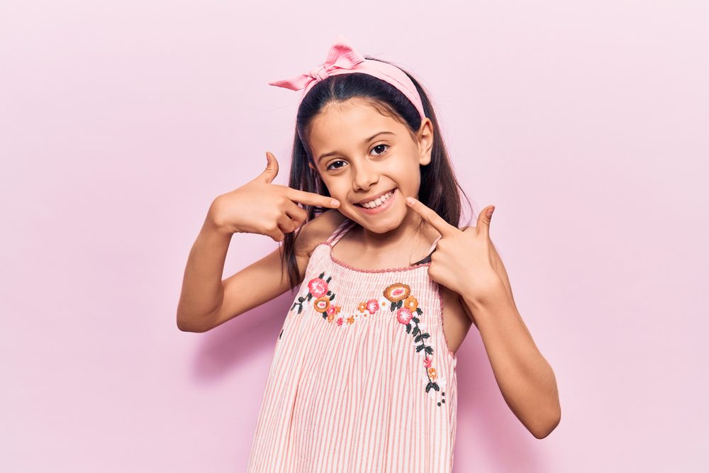 Smiling young girl pointing at her teeth against a pink background